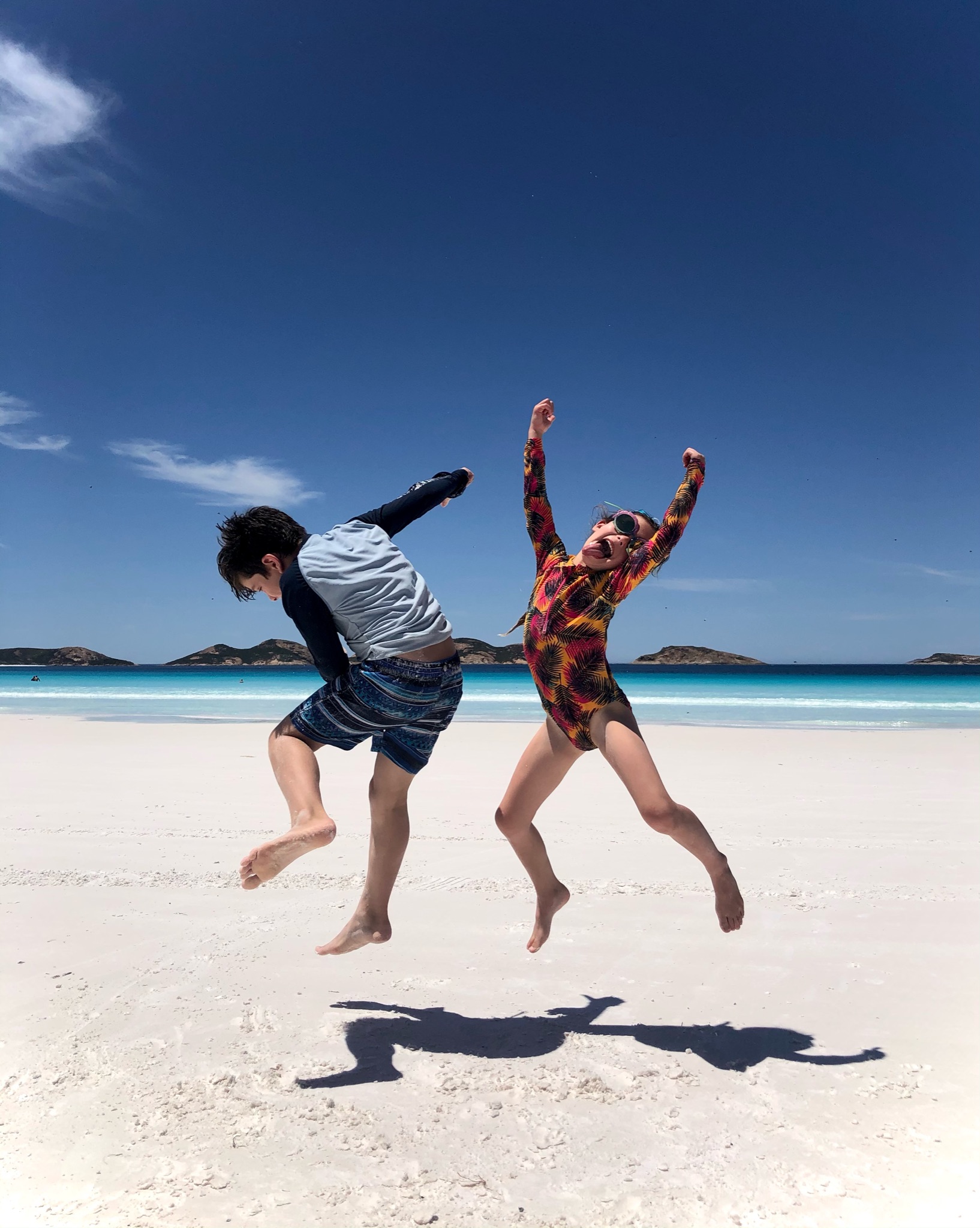 Two children jumping on a beach with very white sand, turquoise water and blue sky